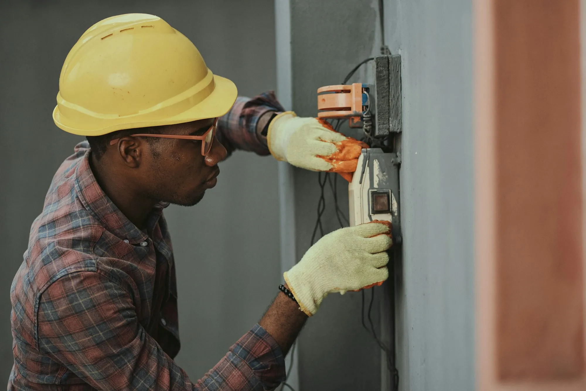 Electrician in a hard hat and gloves working on a wall-mounted panel